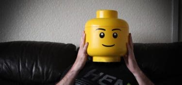 Photo of a person sat on a sofa they are holding their head but their head is a giant yellow lego head.