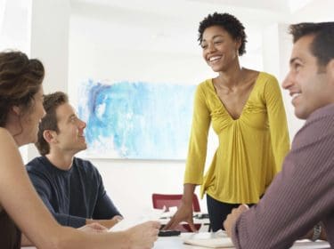 Office with four people sat around a table. There is a lady in a yellow top standing and talking to the group.