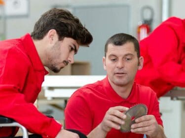Photo of three people in red uniform in a workshop. One person appears to be instructing the other.