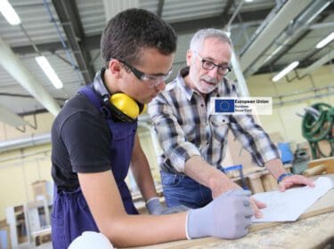 Photo of a younger and older person working together in a warehouse wearing safety gear. The EU social fund logo is at the centre
