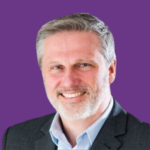 Professional headshot of Jason Colyer smiling, with short grey hair and a trimmed beard, wearing a dark blazer and light blue shirt, against a purple background.