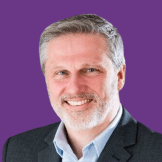 Professional headshot of Jason Colyer smiling, with short grey hair and a trimmed beard, wearing a dark blazer and light blue shirt, against a purple background.