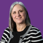 Professional headshot of Jo Lee smiling, with shoulder-length grey hair, wearing a black top and a black-and-white striped cardigan, against a purple background