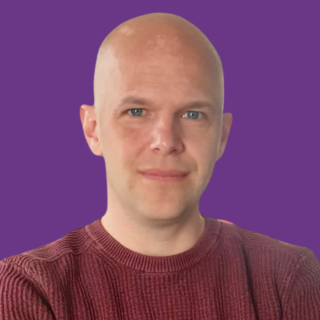 Professional headshot of Joe Ruddock with a shaved head, wearing a burgundy jumper, against a purple background