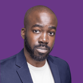 Professional headshot of Joshua Okunlola with a shaved head and beard, wearing a navy blazer over a light-coloured shirt, against a purple background.