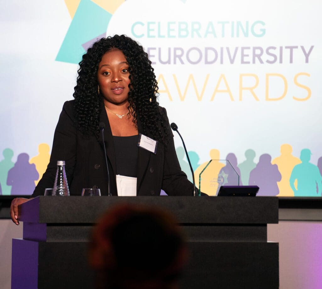 Onyinye accepting her award at the celebrating neurodiversity awards. She is a black lady with long dark hair waering a black suit.