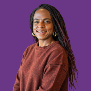 Head-and-shoulders portrait of Claudine Charles smiling with long black braids featuring subtle burgundy highlights, wearing gold hoop earrings and a rust-coloured knitted jumper.
