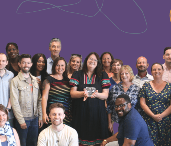 Photo of a diverse group of Genius Within staff all smiling at the camera. The background is dark purple with colourful wavy lines around the edges.