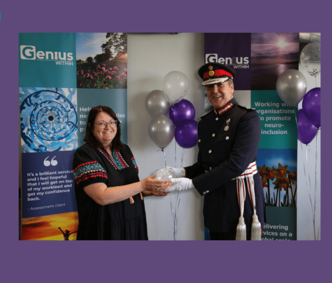 Jacqui Wallis, dressed in a black patterned dress, receives the Kind Award from HM Lord-Lieutenant of East Sussex, Mr Andrew Blackman, who wears a military uniform. They stand in front of balloons and a Genius Within backdrop.