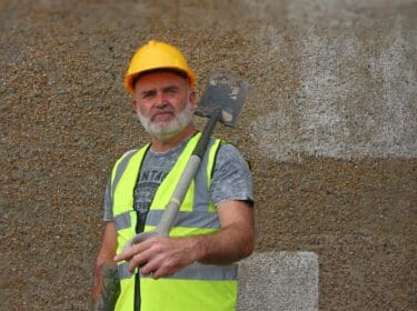 Image of a man in a high vis vest and a protective helmet, holding onto a shovel.
