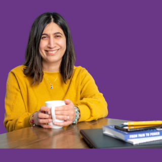 Portrait of Narinder Sheena smiling and holding a mug, wearing a mustard yellow jumper and seated at a table with notebooks, against a purple background.