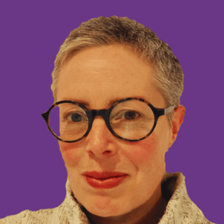 Head-and-shoulders portrait of Morwenna Stewart with short grey hair and round black glasses, looking directly at the camera with a slight smile. They are wearing a light-coloured textured high-neck jumper against a solid purple background.