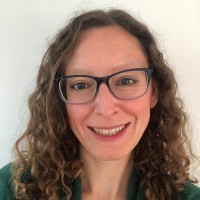 A head and shoulders photo of Helen. Helen has black framed glasses and shoulder length wavy hair. She is smiling and looking directly at the camera