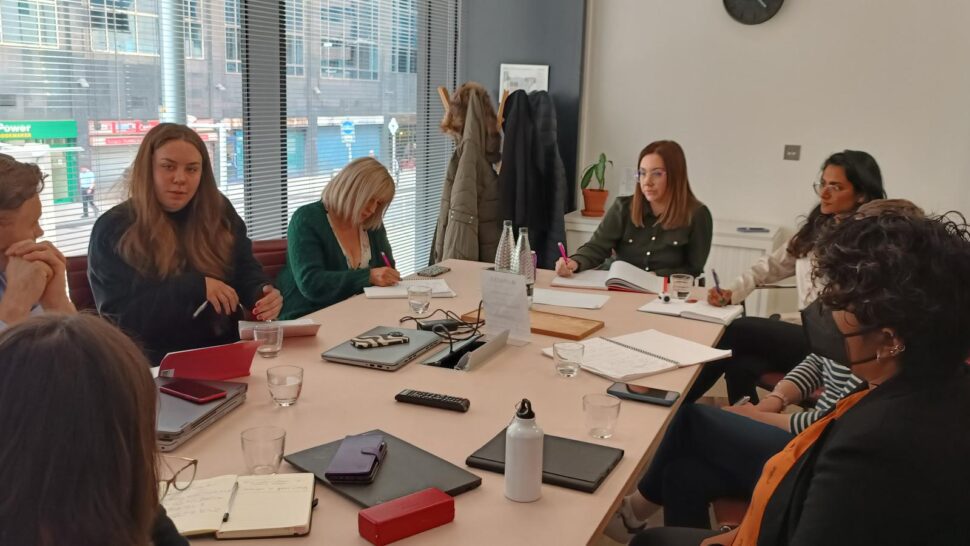 A group of 8 people sat around large table in an office. With a window behind on the left hand side. There is a range of paperwork and laptops on the table.