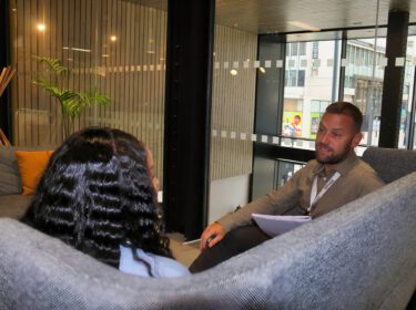 Two people sat on sofa - camera is from behind them. The sofa is grey and there is a glass wall behind them