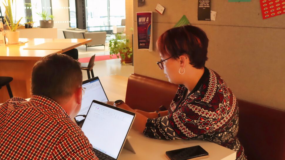 Two people sat at table in office kitchen looking at laptop screen together