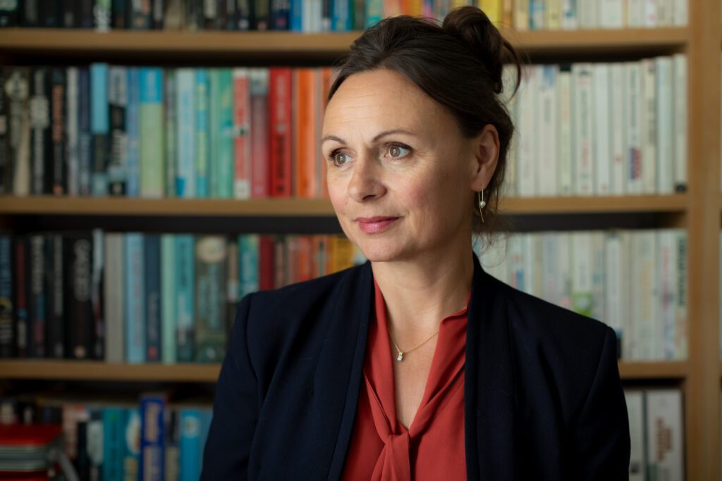 profile photo of Dr Nancy Doyle - she has a library of books in the background. The shot is head and shoulders. She is looking diagonally to her right and wearing a red shirt under a dark blue black jacket.