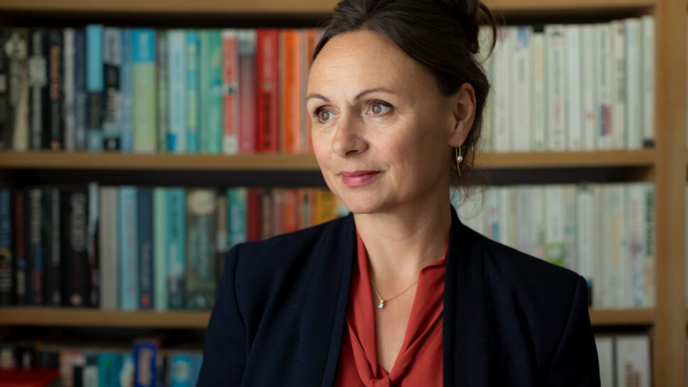 profile photo of Dr Nancy Doyle - she has a library of books in the background. The shot is head and shoulders. She is looking diagonally to her right and wearing a red shirt under a dark blue black jacket.
