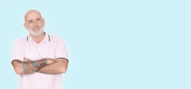 A bald older man with a white beard and colorful tattoo sleeves on one arm stands against a light blue background. He’s wearing a light pink polo shirt and has his arms crossed, looking confidently at the camera with a slight smile.