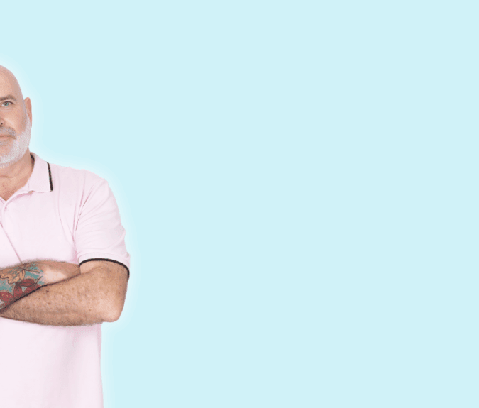 A bald older man with a white beard and colorful tattoo sleeves on one arm stands against a light blue background. He’s wearing a light pink polo shirt and has his arms crossed, looking confidently at the camera with a slight smile.