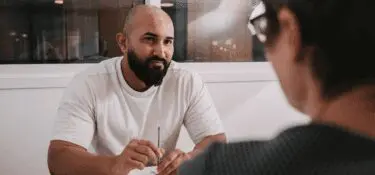 A male wearing white sitting at a desk opposite a female with short hair wearing glasses