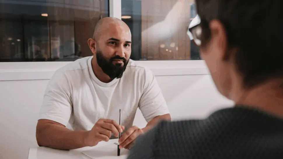 A male wearing white sitting at a desk opposite a female with short hair wearing glasses
