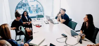 A group of people in a meeting room sitting around a table with laptops and notebooks.