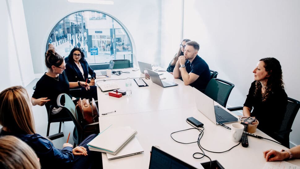 A group of people in a meeting room sitting around a table with laptops and notebooks.