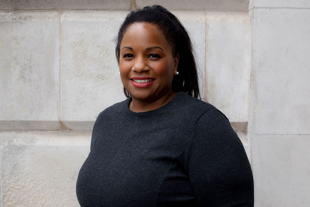 Leah stands outside in front of a grey stone wall. She is looking sideways at the camera and smiling