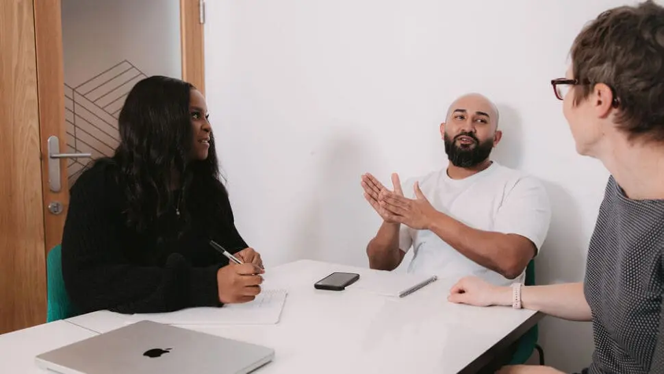 3 people sat at an office table talking. One is the coach, delivery a co-coaching session for a manager and their team member.