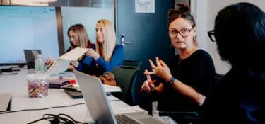 Image shows 4 people sat along one side of an office desk, engaged in conversation with each other. They are engaged in a Leadership Coaching Session.