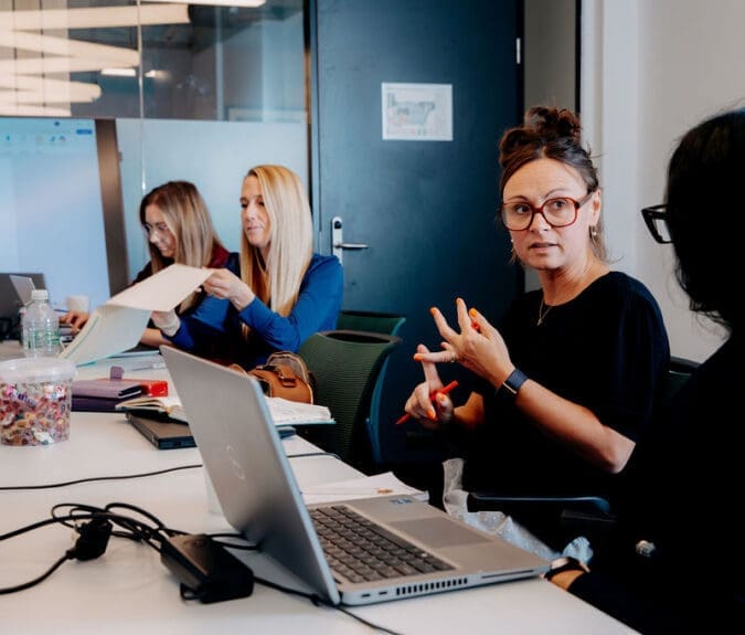 Image shows 4 people sat along one side of an office desk, engaged in conversation with each other. They are engaged in a Leadership Coaching Session.