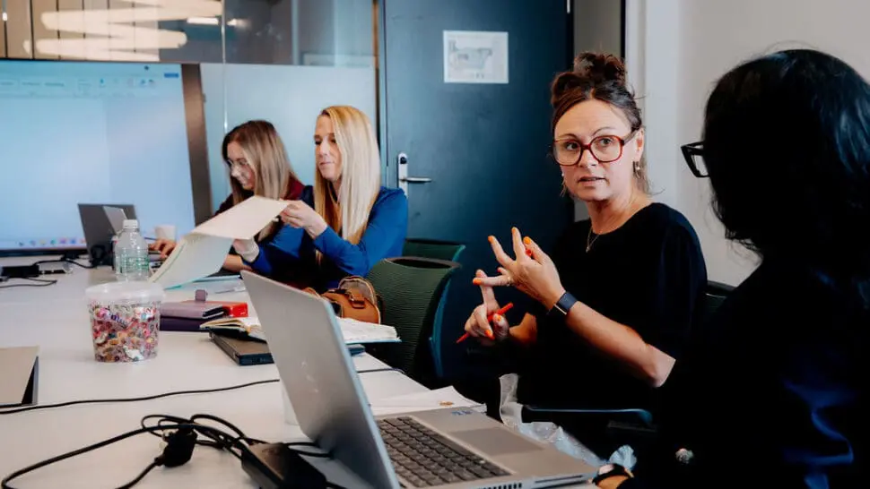 Image shows 4 people sat along one side of an office desk, engaged in conversation with each other. They are engaged in a Leadership Coaching Session.