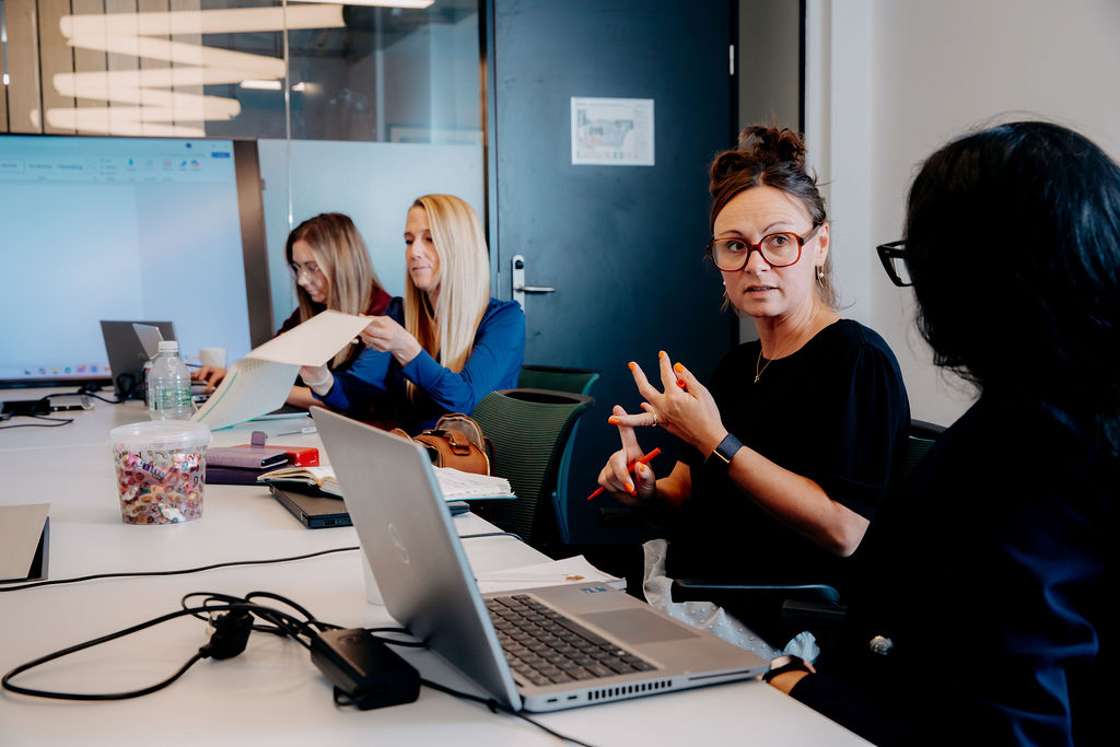 Image shows 4 people sat along one side of an office desk, engaged in conversation with each other. They are engaged in a Leadership Session for Genius Finder 