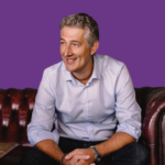 Professional photo of Brendan Dobrowolny sitting on a leather sofa, smiling and wearing a light blue shirt and dark trousers, against a purple background.