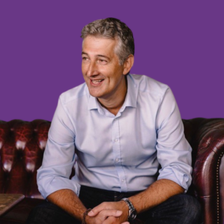 Professional photo of Brendan Dobrowolny sitting on a leather sofa, smiling and wearing a light blue shirt and dark trousers, against a purple background.