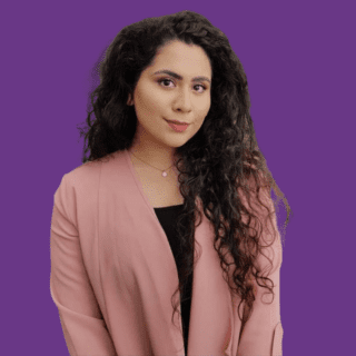 Professional headshot of Aleena Malik against a solid purple background. She is facing the camera with a slight smile and a confident, approachable expression. She has long, dark, curly hair worn loose over one shoulder and is wearing a light pink blazer over a black top, accessorised with a delicate necklace. The portrait is cropped from the waist up.