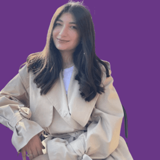 Hazal Seyitoglu seated in a professional portrait against a purple background, wearing a light beige coat and smiling at the camera.