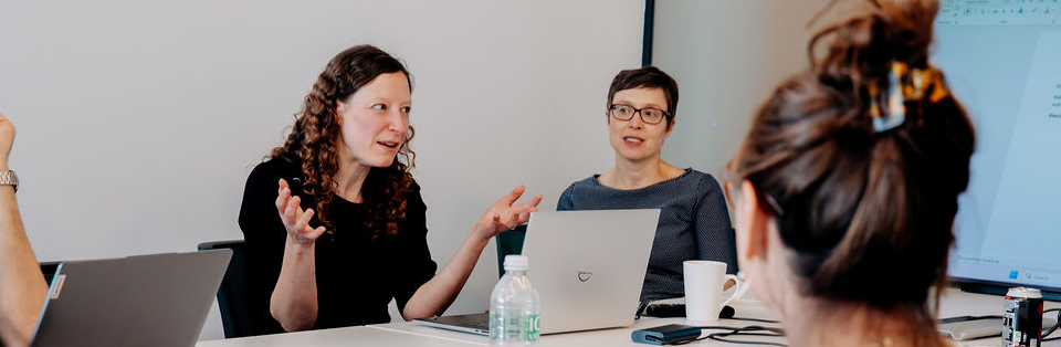 Two people sat at a desk looking back across towards a third person.