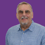 Head-and-shoulders portrait of Malcolm Tydie with short grey hair and glasses, wearing a light blue shirt. He is looking directly at the camera with a gentle smile. The background is dark purple.