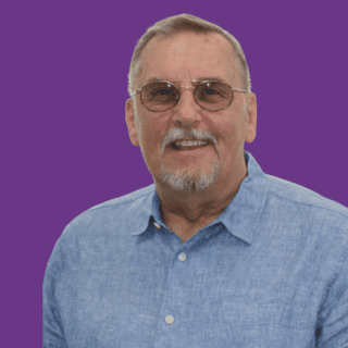 Head-and-shoulders portrait of Malcolm Tydie with short grey hair and glasses, wearing a light blue shirt. He is looking directly at the camera with a gentle smile. The background is dark purple.
