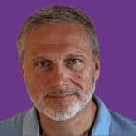 Head-and-shoulders portrait of Jason Colyer, with short grey hair and a neatly trimmed grey beard. He is wearing a light blue collared shirt and looking directly at the camera with a slight smile. The portrait is set against a solid purple background.