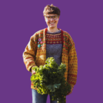 Corinne Cariad smiling and holding a large bunch of fresh kale, wearing glasses and a colourful knitted cardigan against a purple background.