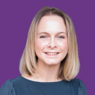 Portrait of Polly smiling at the camera, with shoulder-length blonde hair and wearing a dark blue top against a plain purple background.